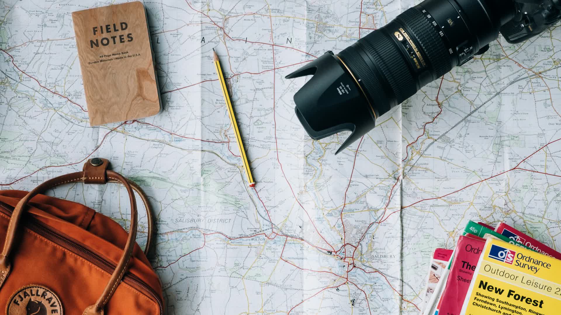 Wide shot: a whole trip-planning desk with a map, camera, notebook and backpack.